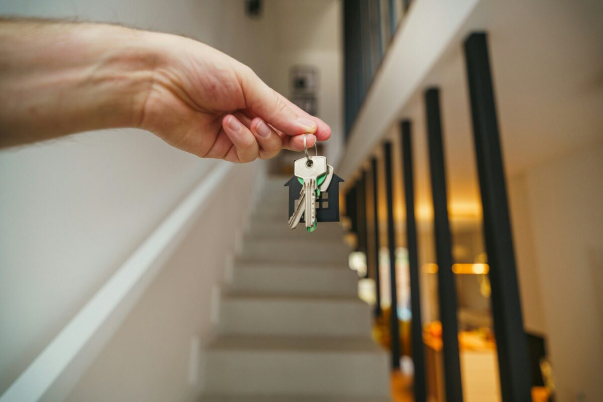 Hand holding a keychain featuring keys and a little house in front of a flight of stairs.