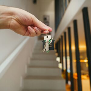 Hand holding a keychain featuring keys and a little house in front of a flight of stairs.